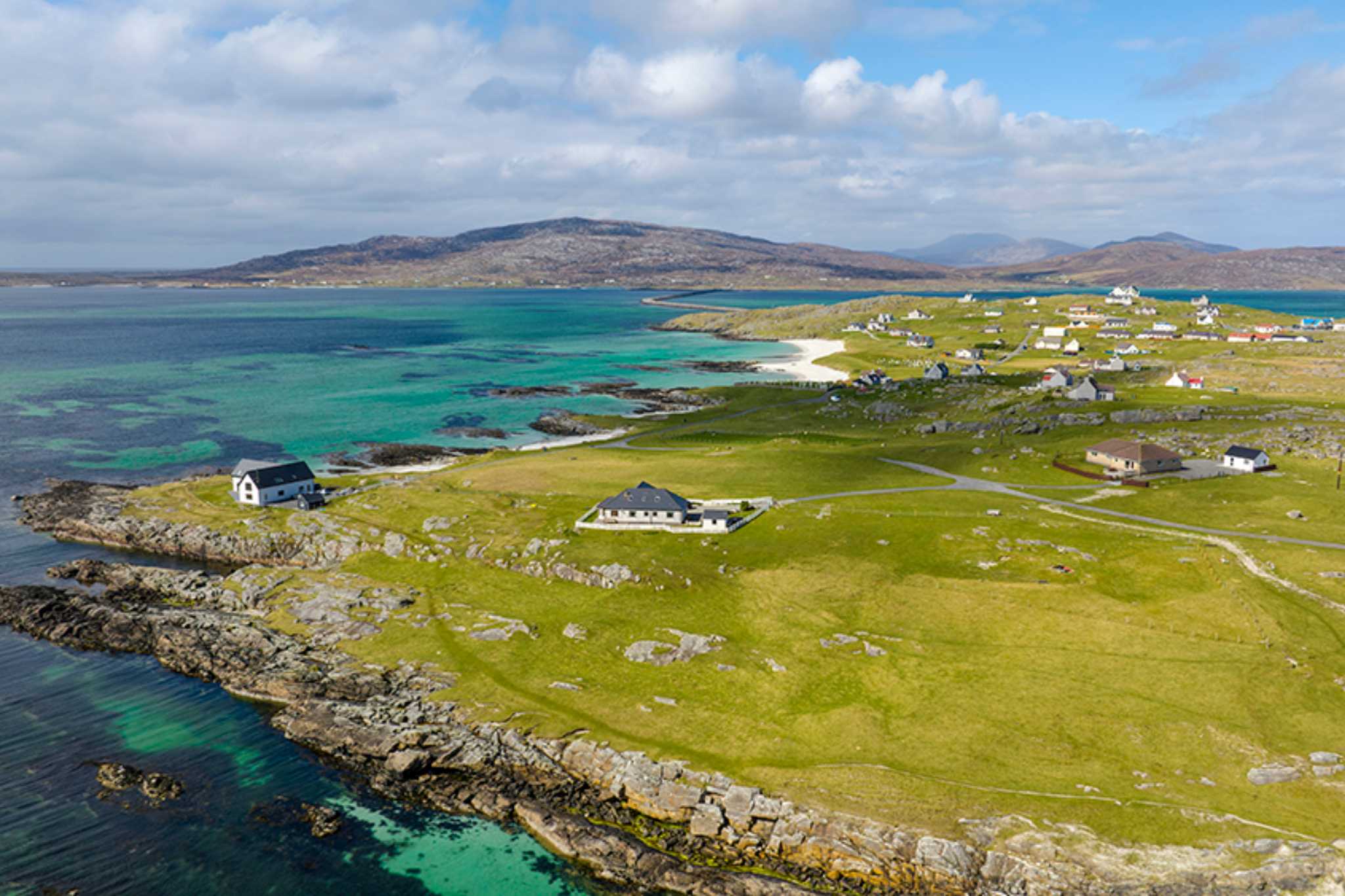 View from the sky of the isle of Eriskay, with turquoise water and white sand beach, and a few houses scattered on the land.