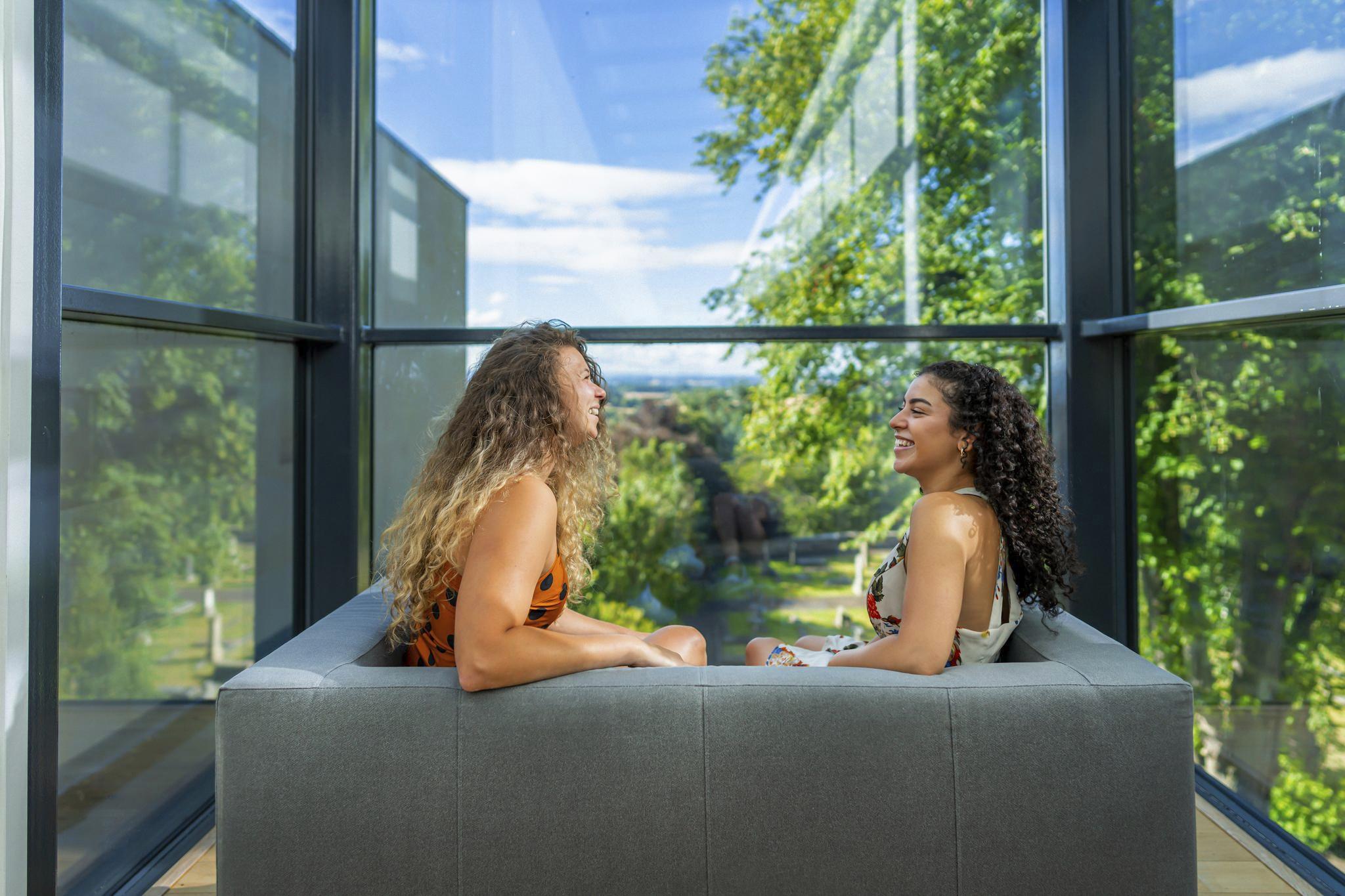 Two visitors sit on a sofa by a large window overlooking park