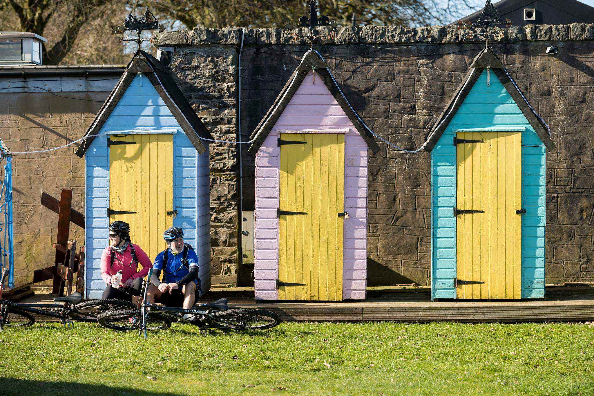 Two cyclists rest in front of three brightly painted beach huts
