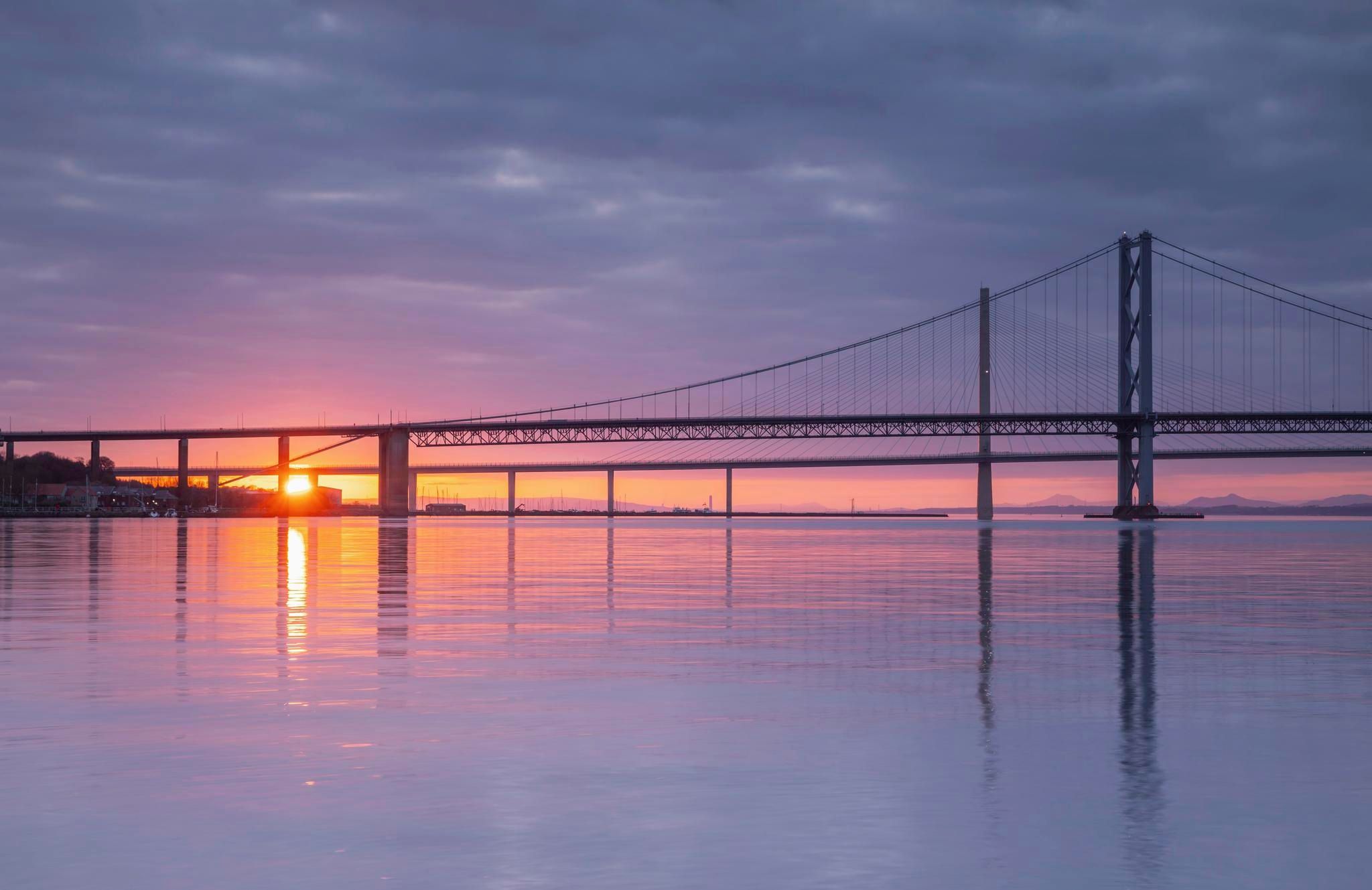 Sun setting over distant hills silhouettes the Forth Road Bridge over calm waters