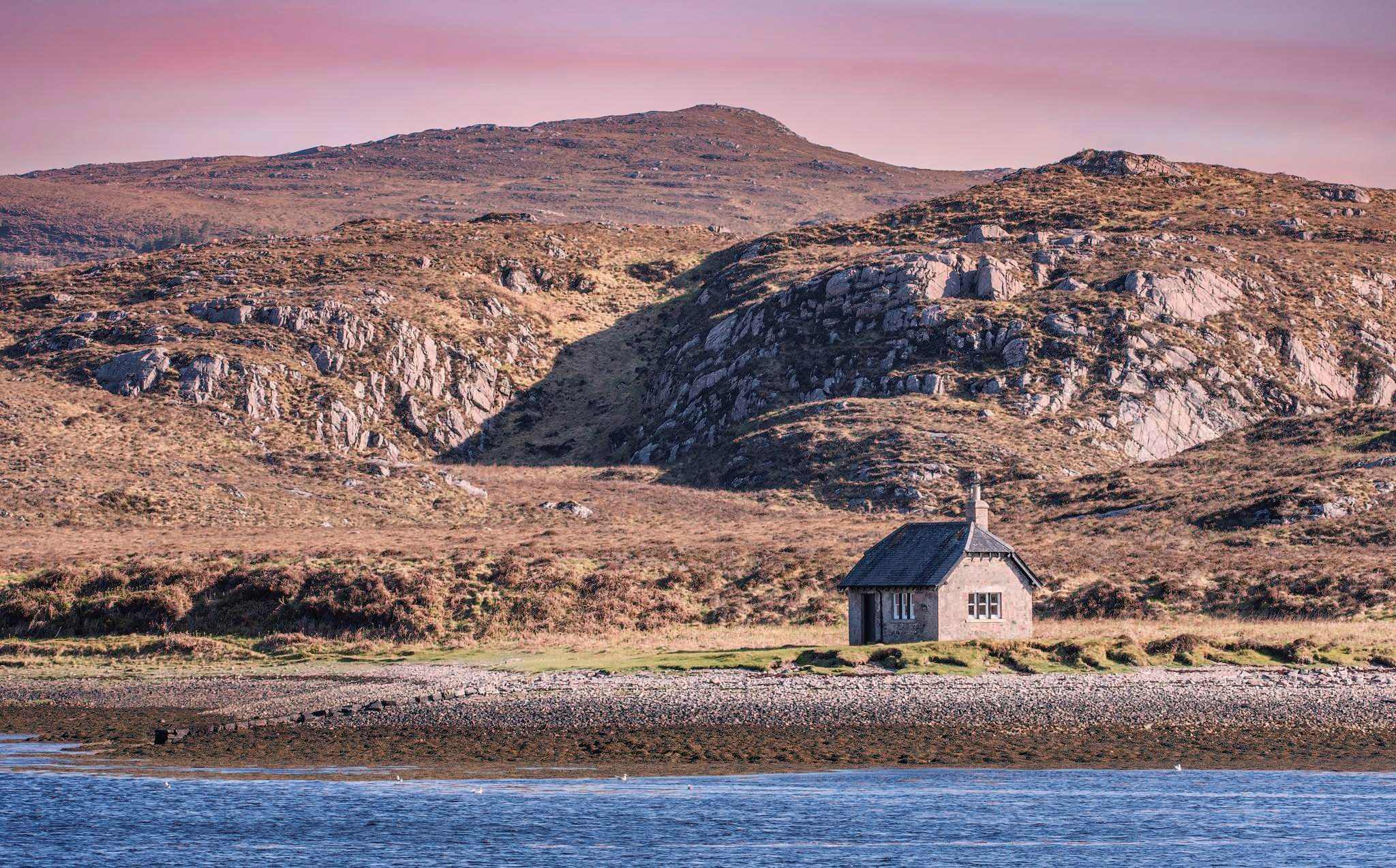 A small dwelling sits on a shoreline blow some rocky hills.