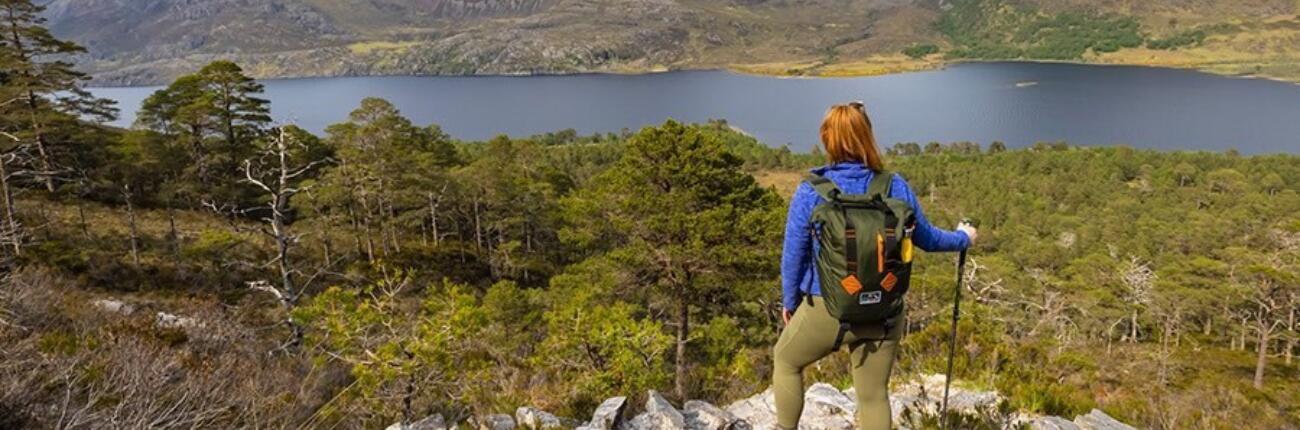 Eine Frau mit Wanderstock blickt von einem felsigen Gipfel auf Bäume, Berge und einen See.