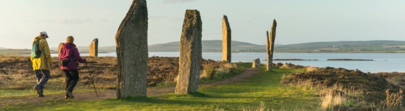 Two people walk over the grass amongst the Ring of Brodgar under a blue sky, the water surrounding the land