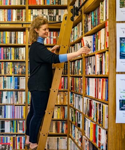 A woman up a ladder against a bookshelf in a busy bookshop