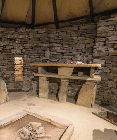 Innenansicht des Nachbaus einer Behausung in Skara Brae mit der Feuerstelle in der Mitte des Raumes.