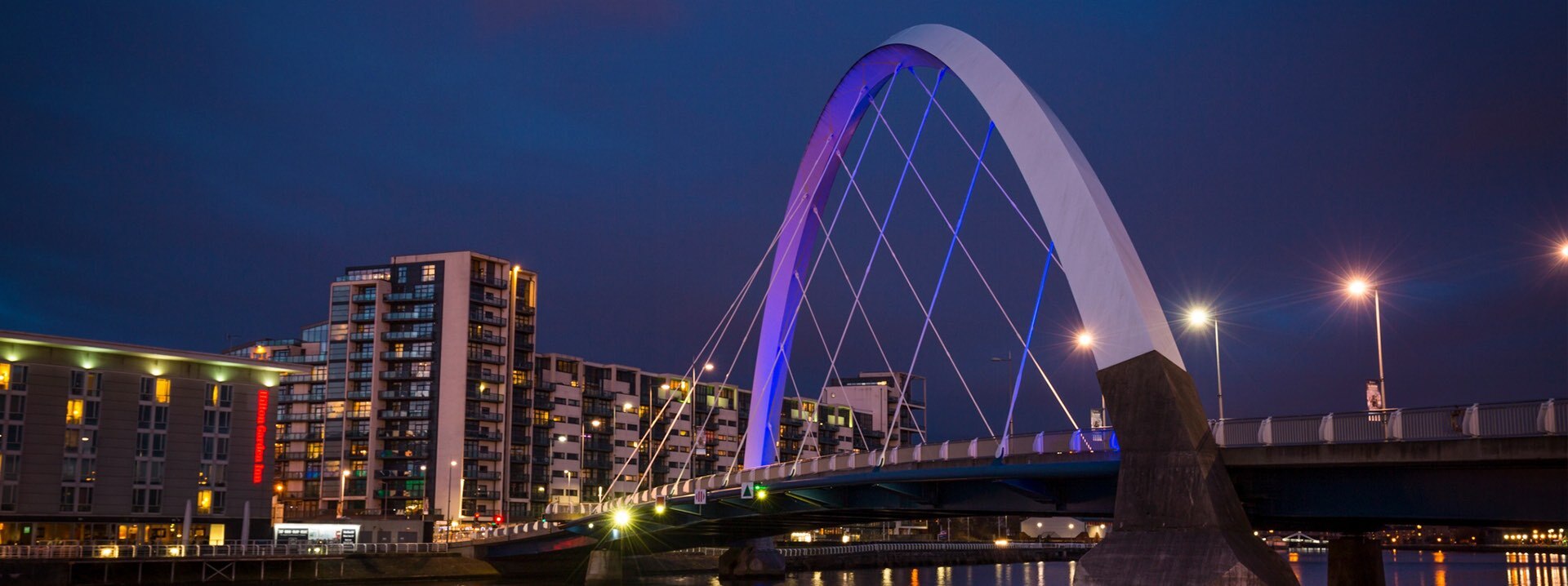 Die beleuchtete Clyde-Arc-Brücke und Gebäude bei Nacht.