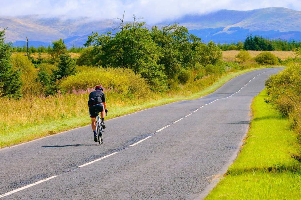 A man cycles along a road, the green grass and bushes on either side, the hills in the background