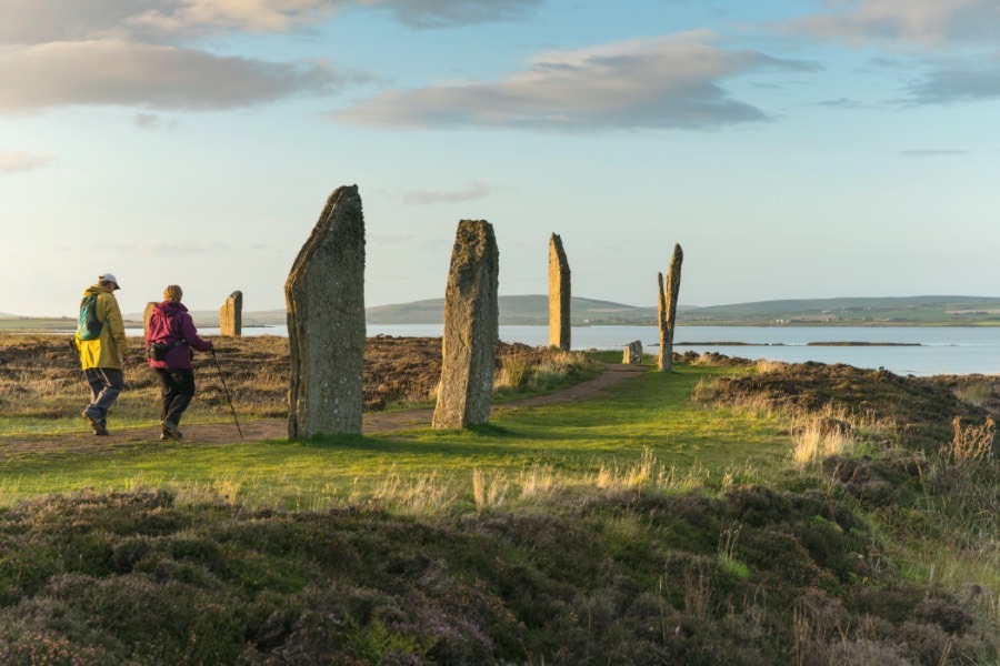 Two people walk over the grass amongst the Ring of Brodgar under a blue sky, the water surrounding the land