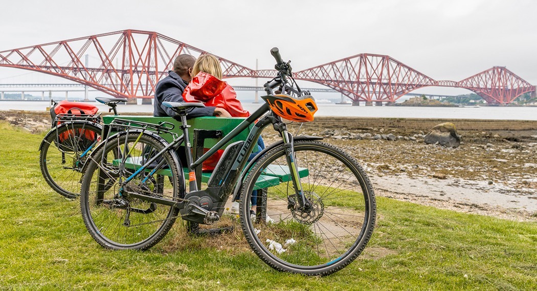 A couple park their bikes to take a break on a bench during a cycle through of the seaside town of South Queensferry, taking in the views of The Forth Bridges (Forth Bridge, The Forth Road Bridge and the Queensferry Crossing
