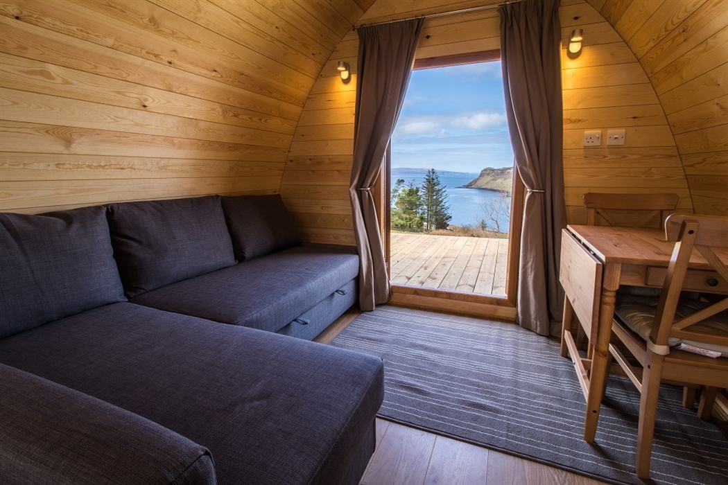 Interior de un pod de camping, con sofás y mesa. Puerta abierta con vistas del mar